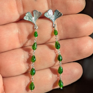 Könnte beinhalten: Silberne Ohrringe mit einem Ginkgoblatt-Design. Jeder Ohrring hat eine Reihe von leuchtend grünen, tropfenförmigen Edelsteinen, die an einer feinen Kette hängen. Die Ohrringe werden vor einem schlichten Hintergrund gezeigt.