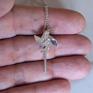 May include: A silver-toned necklace featuring a small, polished pinwheel pendant attached to a delicate chain. The pinwheel has four distinct, angled blades radiating from a central point, suspended from a thin, metallic link chain necklace.