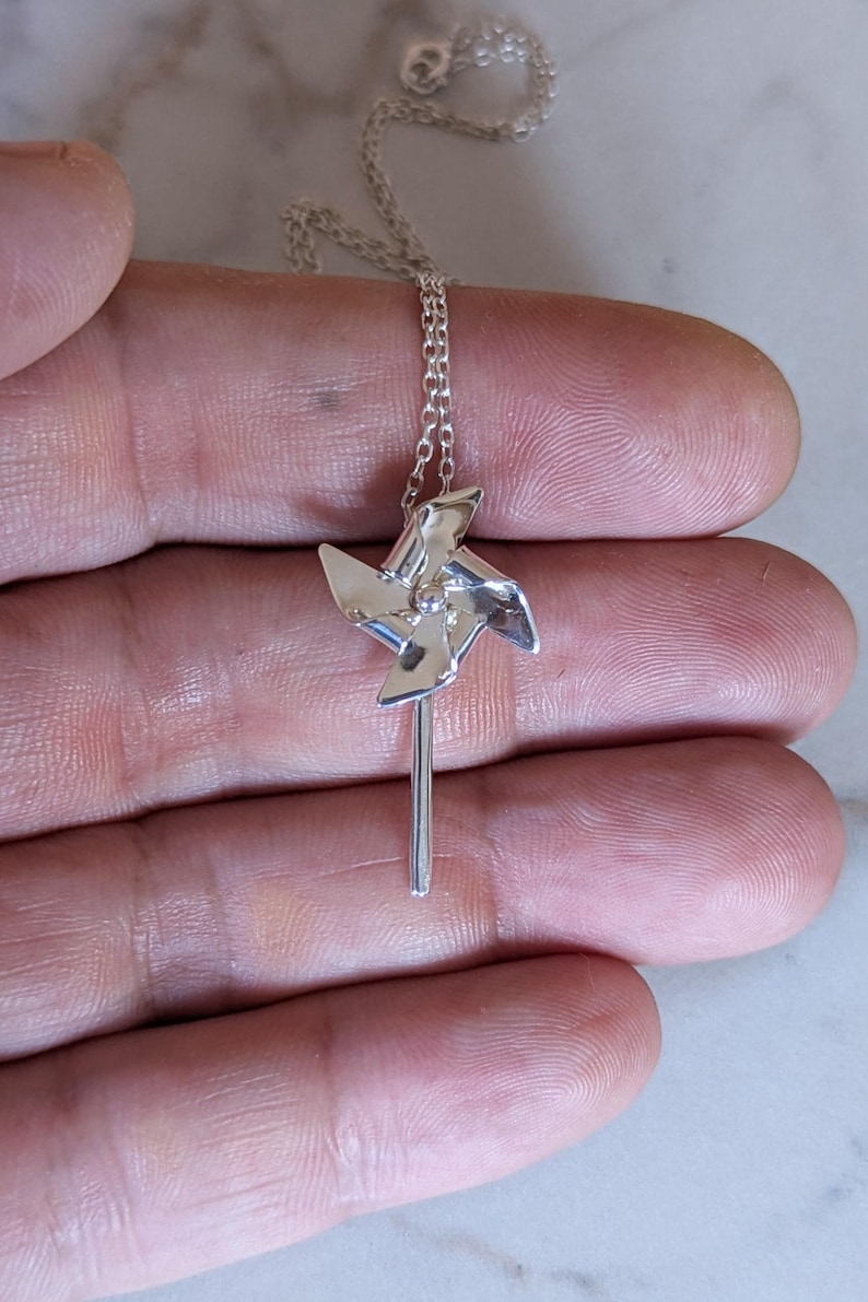 May include: A silver-toned necklace featuring a small, polished pinwheel pendant attached to a delicate chain. The pinwheel has four distinct, angled blades radiating from a central point, suspended from a thin, metallic link chain necklace.