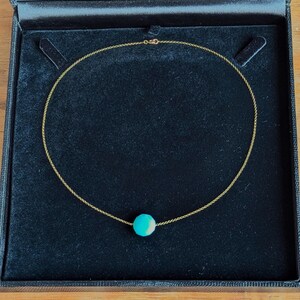 Può includere: Una collana color oro con una singola perla rotonda turchese. La collana è presentata in un cofanetto nero aperto, con interno in velluto. Il cofanetto è appoggiato su una superficie di legno.