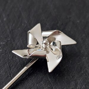 May include: A silver-toned pin featuring a four-bladed pinwheel design. The pinwheel has a polished, reflective surface with curved blades radiating from a central spherical stud. A long, slender metal pin stem extends from the base.