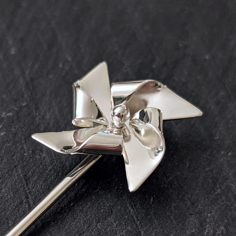 May include: A silver-toned pin featuring a four-bladed pinwheel design. The pinwheel has a polished, reflective surface with curved blades radiating from a central spherical stud. A long, slender metal pin stem extends from the base.