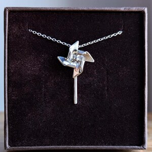 May include: A silver-toned necklace featuring a pendant shaped like a pinwheel with four curved blades attached to a vertical stem. The pendant is suspended from a delicate silver-toned chain, showcasing a polished metallic finish throughout.