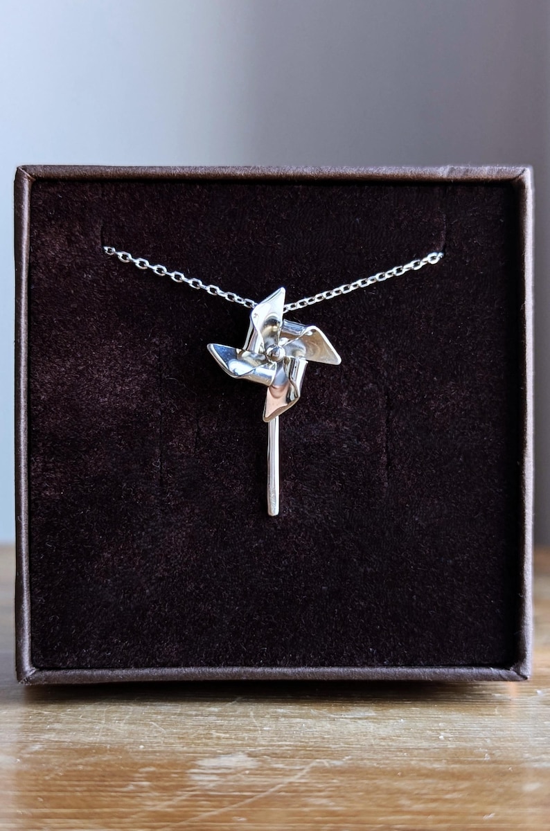 May include: A silver-toned necklace featuring a pendant shaped like a pinwheel with four curved blades attached to a vertical stem. The pendant is suspended from a delicate silver-toned chain, showcasing a polished metallic finish throughout.
