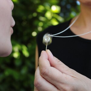 May include: A silver chain necklace featuring a small, clear, spherical pendant with a swirling white interior design. The pendant is held between fingers, showcasing its transparent glass-like texture and the delicate metallic chain.