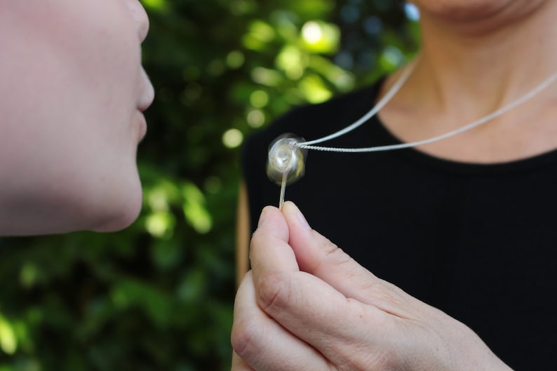May include: A silver chain necklace featuring a small, clear, spherical pendant with a swirling white interior design. The pendant is held between fingers, showcasing its transparent glass-like texture and the delicate metallic chain.