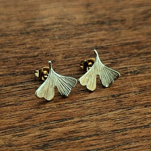 Peut inclure: Une paire de boucles d'oreilles à clous en forme de feuilles de ginkgo dorées. Chaque boucle présente une silhouette en éventail avec des lignes gravées rayonnantes imitant les nervures naturelles, dotée d'une tige et d'un fermoir papillon.