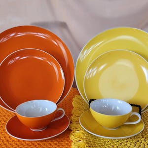 Puede incluir: Un juego de vajilla de cerámica naranja y amarilla. Cada juego incluye un plato grande, un plato más pequeño, un platillo y una taza de té. Las tazas y platillos son naranjas y amarillos, con interiores blancos. Los platos tienen bordes blancos.