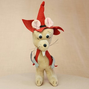 Könnte beinhalten: Ein Plüschtier in Form einer Maus, das einen roten spitzen Hut mit rosa Ohren trägt. Die Maus hat einen beigefarbenen Körper, schwarze Augen und eine schwarze Nase. Das Spielzeug steht auf einer hellbraunen Oberfläche.