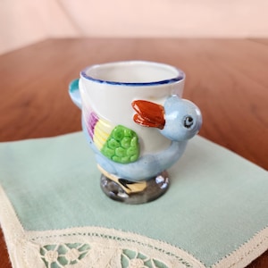 May include: A blue and white ceramic egg cup shaped like a duck with a colourful design. The duck has a yellow beak and orange feet.
