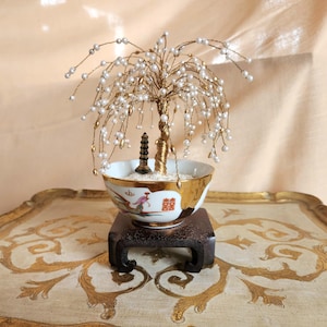 May include: A decorative bonsai tree with a gold-colored wire trunk and branches adorned with white pearls. The tree sits in a white and gold bowl with a small pagoda and is displayed on a wooden stand. The bowl has a red and gold design.