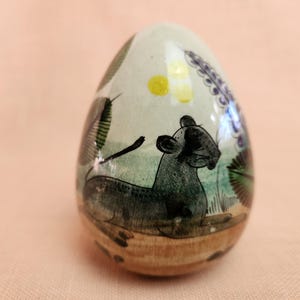 May include: A ceramic egg-shaped ornament with a hand-painted design of a black and green animal, possibly a jaguar, lying down in a green and brown landscape. The background features a yellow sun and purple flowers.