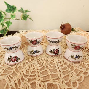 May include: Four white ceramic egg cups with floral designs in shades of pink, blue, and green. The egg cups are stacked on a white crocheted doily.