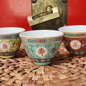 May include: Three small, decorative porcelain tea cups with white interiors. Each cup features a colorful, ornate design with Chinese characters and floral patterns. The cups are set on a woven mat against a red background.