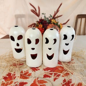 May include: Four white, ghost-shaped candle holders with carved-out eyes and mouths, arranged on a table with fall-themed decorations. The candle holders have a smooth finish and a hollow interior. The table is covered with a leaf-patterned cloth.