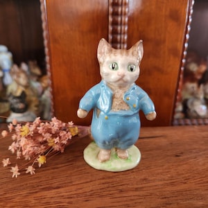 Puede incluir: Una figurita de porcelana azul y blanca de un gato con una chaqueta y pantalones azules. La figurita está de pie sobre una base verde.