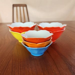 Puede incluir: Un conjunto de cuencos coloridos con forma de flor en tonos rojos, amarillos, naranjas y azules. Los cuencos tienen un interior blanco y están apilados sobre una superficie de madera. Una silla de madera está en el fondo.