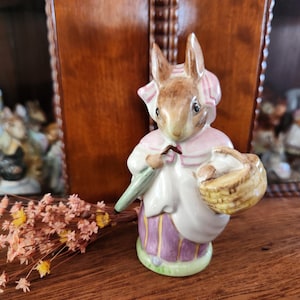 Pode incluir: Uma figurinha de porcelana de um coelho vestindo um vestido rosa e um chapéu verde, segurando uma cesta e um guarda-chuva verde. A figurinha está em pé sobre uma superfície de madeira.