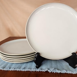 Puede incluir: Un conjunto de cuatro platos de cena de cerámica blanca con un borde ligeramente elevado. Los platos están apilados sobre un mantel individual azul y blanco.