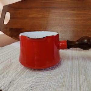 May include: A small, red enamel saucepan with a dark wooden handle. The interior is white, and the rim is black. The saucepan sits on a white cloth, with a wooden tray in the background. A vintage kitchenware item.