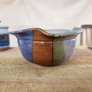 May include: A handmade ceramic mixing bowl with a spout, featuring sections of blue, brown, and green. The bowl sits on a wooden surface, with a small jar with a lid in the background. This unique bowl is perfect for kitchen use.