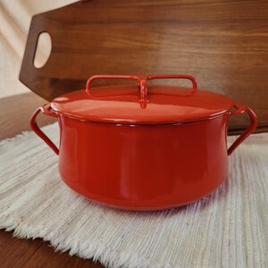 May include: A bright red enamel pot with a matching lid. The pot has two side handles and a handle on the lid. The pot is sitting on a white, textured placemat. A wooden cutting board is in the background.