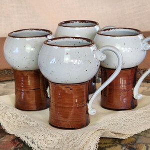 Puede incluir: Cuatro tazas de cerámica hechas a mano. Cada taza tiene un cuerpo blanco moteado, una base marrón y un asa curva. Las tazas están dispuestas sobre una bandeja decorativa con un mantel de encaje.