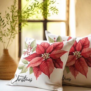 Può includere: Due cuscini quadrati a tema natalizio. Ogni cuscino presenta un grande e dettagliato fiore di poinsettia nei toni del rosso e del verde su uno sfondo color crema. Le parole "Merry Christmas" sono scritte in corsivo sul davanti.