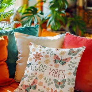 Può includere: Un cuscino decorativo con un motivo floreale e farfalle, con il testo "GOOD VIBES". Il cuscino ha uno sfondo bianco con accenti rosa, verdi e gialli. Sono visibili altri cuscini nei toni dell'arancione, del verde acqua e del crema.