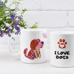 Può includere: Due tazze in ceramica bianca con disegni a tema cani. Una tazza presenta un cane dei cartoni animati con un naso rosa e accenti floreali. L'altra tazza ha un'impronta di zampa con un cuore e il testo "I LOVE DOGS".