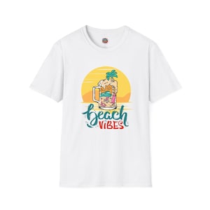 Può includere: T-shirt bianca con una grafica colorata. Il design mostra un boccale di birra con una scena di spiaggia, una palma e le parole "beach vibes" in un carattere script. Il design include un sole giallo e onde.
