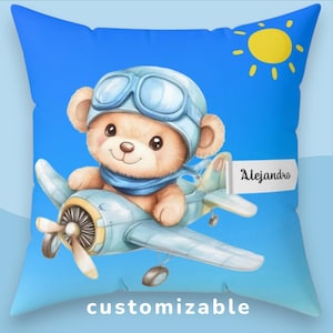 Può includere: Un cuscino quadrato con sfondo blu. Presenta un orsacchiotto dei cartoni animati con casco e sciarpa da pilota, che vola su un aereo azzurro. Il cuscino ha il nome "Alejandro" e la parola "customizable". Un sole giallo è nell'angolo in alto a destra.