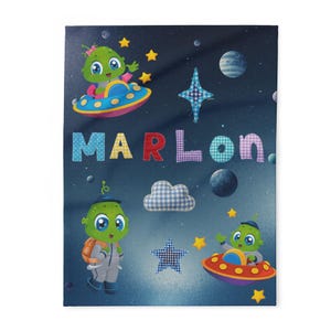 Può includere: Una coperta colorata a tema spaziale, con alieni dei cartoni animati in navicelle spaziali, pianeti e stelle. Il nome "MARLON" è scritto a grandi lettere decorate. Lo sfondo è blu scuro con un effetto maculato.