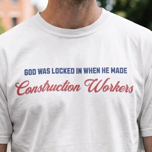 May include: A white short-sleeved t-shirt featuring the text "GOD WAS LOCKED IN WHEN HE MADE Construction Workers" printed on the chest. The first line of text is in blue sans-serif font, and the second line is in a red cursive script font.