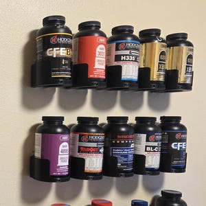 May include: Assortment of black powder containers with various labels, including "H110", "HS-6", and "No. 7", mounted on a wall. The containers are held in black plastic holders.