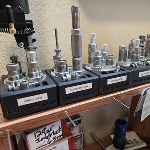 Pode incluir: Um conjunto de matrizes de recarga, incluindo 9MM Luger, 223 Remington e 300 Blackout, exibidas em uma prateleira de madeira. As matrizes prateadas e pretas estão organizadas em suportes pretos, rotulados com a marca CMAprecision.