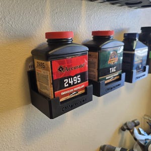 May include: Black containers of smokeless propellant are displayed on a wall. The containers have red lids and labels with text including "Accurate 2495" and "TAC Smokeless Powder." They are held in black wall-mounted holders.