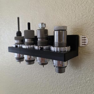 Peut inclure: Un organisateur mural noir contenant cinq matrices de rechargement métalliques. Les matrices sont cylindriques avec divers modèles supérieurs, dont un écrou hexagonal et un capuchon moleté. L'organisateur a une bande adhésive avec des indicateurs de flèches.