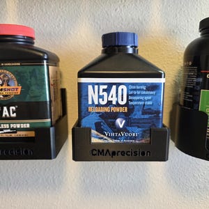 May include: Three black containers of reloading powder are displayed. The containers have labels with text including "TAC Smokeless Powder", "N540 Reloading Powder", and "StaBALL 6.5 Smokeless". Each container is held in a black holder with the "CMAprecision" logo.