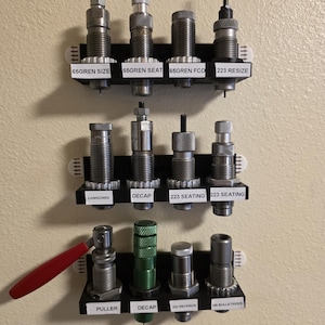 May include: A collection of reloading dies, organized on a wall-mounted black rack. The dies are silver and black, with labels indicating their function, such as "65GREN SIZE" and "223 RESIZE". A red handle is visible on one of the tools.