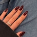 May include: Close-up of a hand with long, almond-shaped nails painted a deep burgundy red. The nails are manicured and have a glossy finish. A gold ring with small stones is worn on the ring finger.