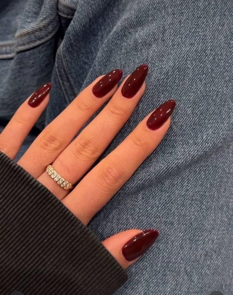 May include: Close-up of a hand with long, almond-shaped nails painted a deep burgundy red. The nails are manicured and have a glossy finish. A gold ring with small stones is worn on the ring finger.