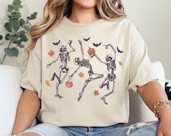 May include: Beige t-shirt with a Halloween design. The graphic shows three dancing skeletons, pumpkins, bats, and autumn leaves. The skeletons are depicted in motion, holding pumpkins, and surrounded by bats and falling leaves.
