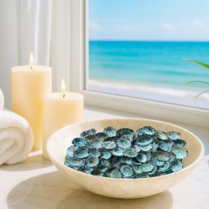 May include: A spa-themed still life featuring rolled white towels, lit candles, and a bowl filled with blue decorative shells. A window offers a view of the ocean and a potted plant.