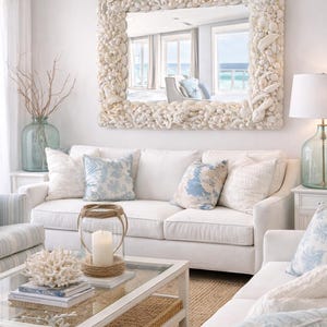 May include: A bright, airy living room with white sofas and blue patterned pillows. A large mirror framed with seashells hangs above the sofa. A glass coffee table displays decorative coral and books, creating a coastal theme.