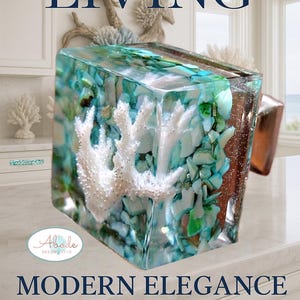 Puede incluir: Un cubo decorativo de resina con un diseño de coral blanco, piedras turquesas y verdes, y el texto "COASTAL LIVING" y "MODERN ELEGANCE". El cubo está sobre una superficie blanca.