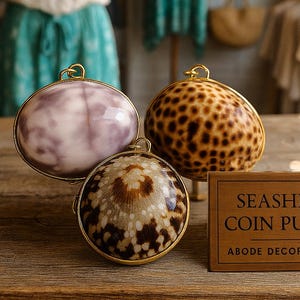May include: Three seashell coin purses with gold frames sit on a wooden surface. The purses feature distinctive patterns: one is lavender, one is spotted, and the other is brown and white. A wooden sign reads "Seashell Coin Purse" and "Abode Decor Gifts."