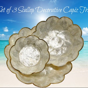 May include: Set of three white scallop-shaped decorative trays with gold trim. The trays are made of capiz shells and are nested together. The trays are photographed against a beach background.