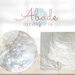 May include: A circular and a square coaster featuring a shimmering, iridescent mother-of-pearl finish with natural shell textures. Above the coasters, the text Abode DECOR GIFTS is displayed in a rose gold script and block font.