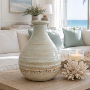 May include: A ceramic vase with a bulbous base and a narrow neck, featuring a crackled glaze finish in shades of light blue and beige. The surface has horizontal ridges and a textured, uneven bottom edge with a natural, earthy appearance.